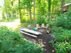 A forested outdoor area with a picnic table, chairs, and a stone fireplace.