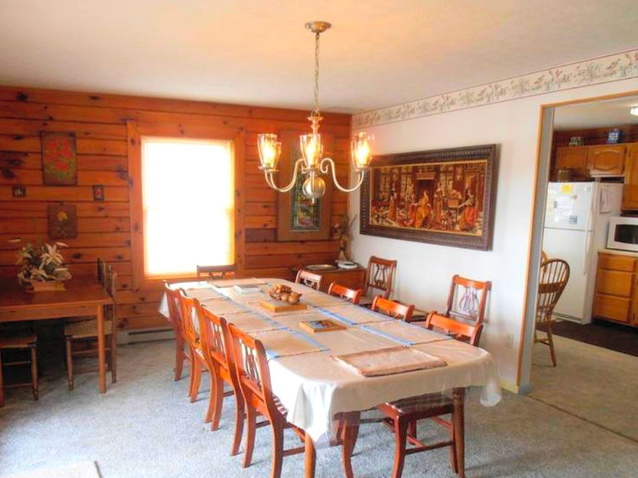 A dining room with a long table, wooden chairs, a chandelier, and a kitchen visible through an open doorway.