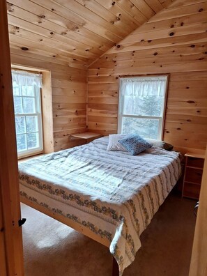 Une chambre dans une cabane en bois, avec un lit, deux fenêtres et une petite table de chevet.