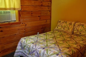 A wooden-paneled room with a bed featuring a floral quilt, a window with a yellow curtain, and a wall-mounted air conditioning unit. - Rustic Family-Friendly Log Cabin in the Smoky Mountains, North Carolina (Cherokee)