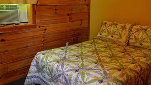 A wooden-paneled room with a bed featuring a floral quilt, a window with a yellow curtain, and a wall-mounted air conditioning unit. - Rustic Family-Friendly Log Cabin in the Smoky Mountains, North Carolina (Cherokee)