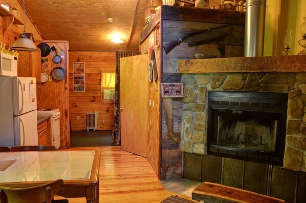 A cozy cabin interior with a stone fireplace, wooden walls, a dining area, and a kitchen with a refrigerator and stove.