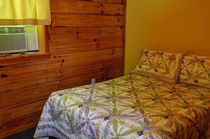 A wooden-paneled room with a bed featuring a floral quilt, a window with a yellow curtain, and a wall-mounted air conditioning unit.
