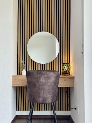 Ein Stuhl, der zu einer Wand mit vertikaler Holzlamellenstruktur zeigt, ein runder Spiegel, ein kleiner weißer Vasen und eine geometrisch gemusterte Lampe auf einem Holzregal.
