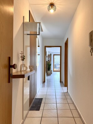 Um corredor com porta de madeira, prateleira com planta, luminária e vista para área externa.