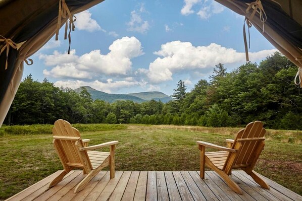 A wooden deck with two Adirondack chairs facing a grassy field and forested mountains under a partly cloudy sky.