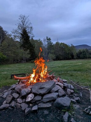 A campfire in a natural setting with trees and a grassy field in the background.