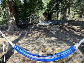 A hammock tied between two trees, a small wooden cabin, and a stone-paved area. - Scenic Off-Grid Campsite Overlooking Crystal Clear Lake in Sagle, Idaho (Sagle)