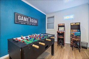 Ein Raum mit einem Kicker, Spielautomaten und einem leuchtenden Neon-Schild mit der Aufschrift 'GAME ROOM'.