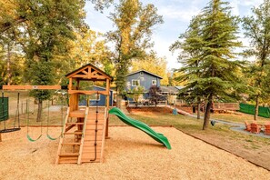 Un parque infantil con un tobogán de madera y columpios, rodeado de árboles y una casa.