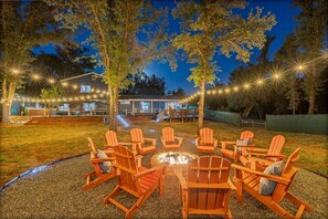 Un espacio al aire libre con sillas Adirondack y una chimenea, rodeado de árboles y una casa.