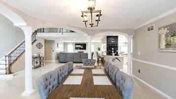 A spacious living area with a wooden dining table, grey upholstered sofas, a kitchen area, and a staircase.