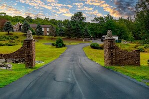 A paved road leading to a stone gate with lion statues, surrounded by greenery and residential buildings. - Modern Villa with Sauna, Pool & Panoramic Views in Bushkill, Pennsylvania (Bushkill)