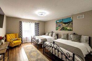 A bedroom with two beds, a yellow armchair, a wooden table, and a cityscape painting.