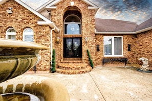 A brick house with a large arched entrance, a stone fountain, and a well-maintained lawn.