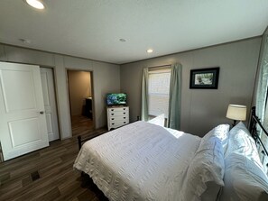 Un dormitorio con cama, cómoda, televisor, ventana con cortinas y puerta.