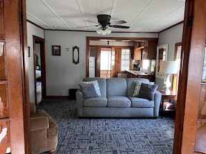 Ein Wohnzimmer mit grauem Sofa, Holzboden, Deckenventilator und einer Tür zu einem anderen Raum.