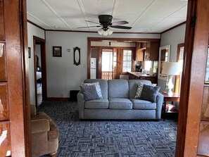 A living room with a gray sofa, wooden floor, ceiling fan, and a door leading to another room. - Spacious Modern Cabin with a Fire Pit near Apostle Islands in Wisconsin (Ashland)