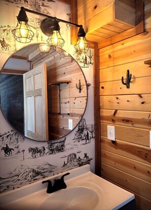 Ein Holzblockhaus-Interieur mit einem runden Spiegel, Holzvertäfelung an den Wänden und einer Deckenleuchte über dem Waschbecken.