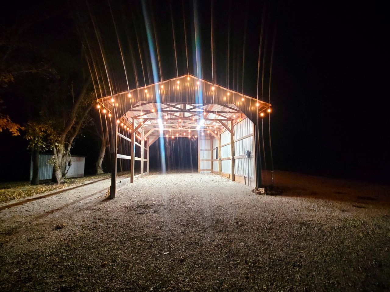 A covered outdoor area with a gravel floor, surrounded by trees and a building in the background.