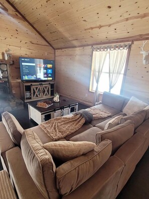 A cozy cabin interior with a wooden ceiling, a television on a wooden stand, a sofa with a blanket, and a window with curtains. - Peaceful Woodland Cabin with Balcony & Starry Nights in Steelville, Missouri (Steelville)