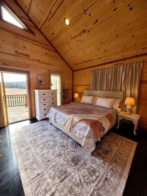 Ein Schlafzimmer in einer Holzhütte mit einem Bett, zwei Nachttischen mit Lampen, einer Kommode und Blick ins Freie durch eine Tür.