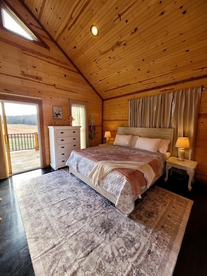 Ein Schlafzimmer in einer Holzhütte mit einem Bett, zwei Nachttischen mit Lampen, einer Kommode und Blick ins Freie durch eine Tür.
