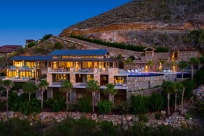 A multi-story stone building with a swimming pool, surrounded by palm trees and a hilly landscape.