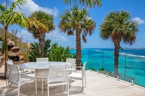 Um pátio com uma mesa redonda e cadeiras, com vista para um mar azul-turquesa e palmeiras.
