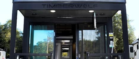 Una caravana recreativa negra con el logo 'Timberwolf', una puerta corredera de acceso y una pequeña zona de porche con asientos.