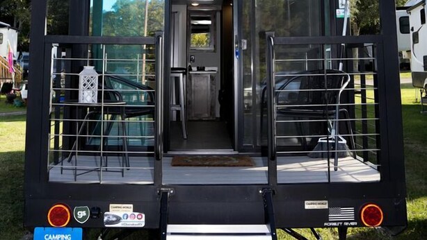 Una caravana recreativa negra con el logo 'Timberwolf', una puerta corredera de acceso y una pequeña zona de porche con asientos.