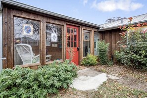 A wooden house with a red door, large windows, and a small garden with a chair and plants. - Riverbank Escape for Families and Friends with Ping Pong, Mrs. Pac-Man, and Cozy Den in Kerrville, Texas (Kerrville)