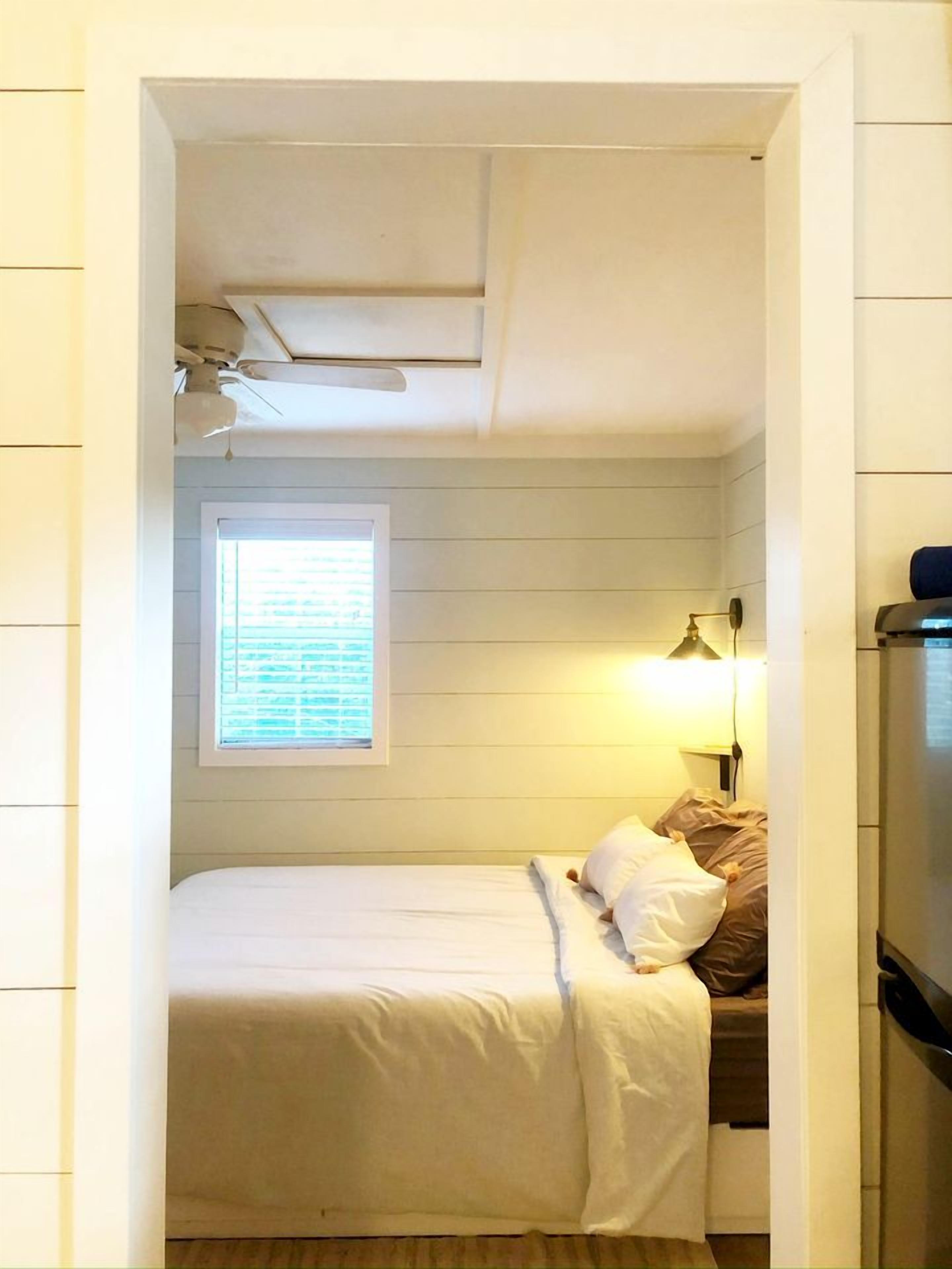 A small bedroom with a bed, a window with blinds, a ceiling fan, and a nightstand with a lamp.