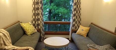 Una sala de estar acogedora con dos sofás, una mesa de centro redonda y vistas a árboles a través de una ventana.