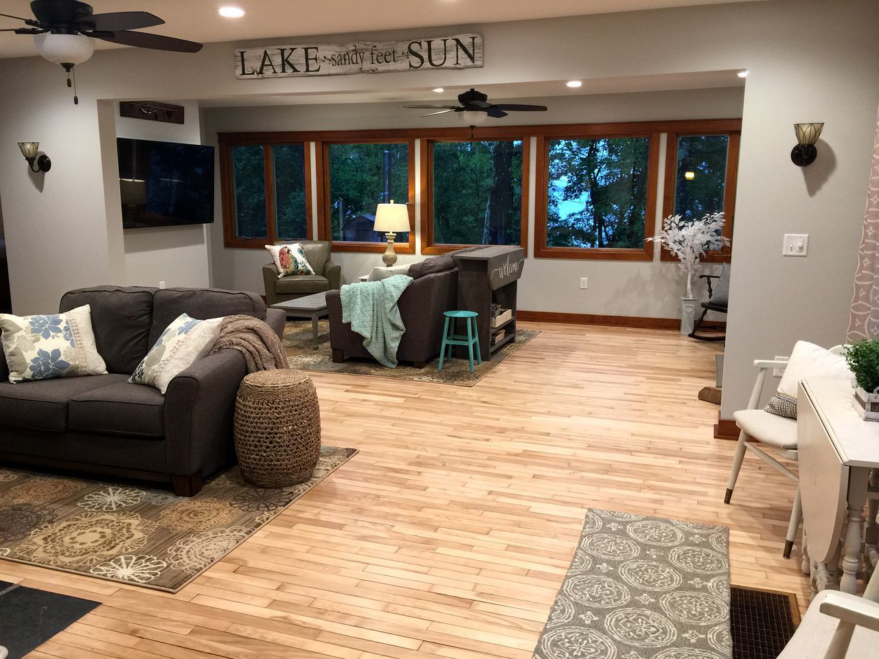 A spacious living area with a sofa, a coffee table, and a dining table. There is a ceiling fan, a TV mounted on the wall, and large windows offering a view of the outdoors.