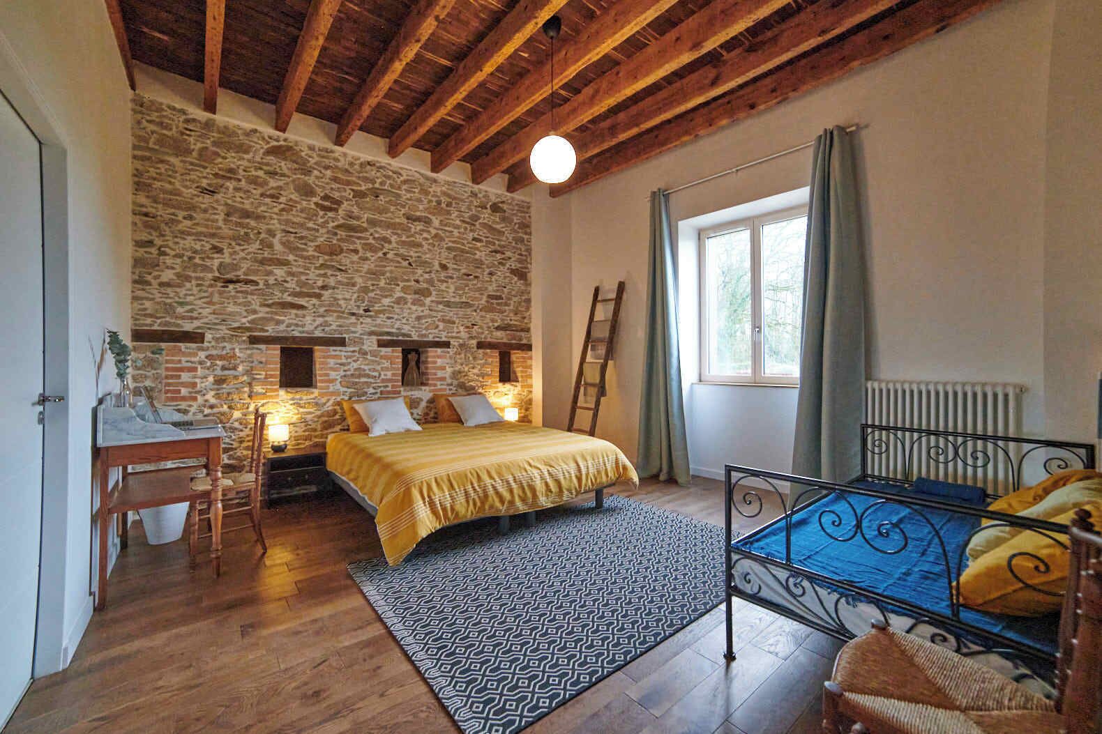 A bedroom with a stone wall, wooden ceiling beams, a bed with a yellow bedspread, a desk with a chair, and a window with curtains.