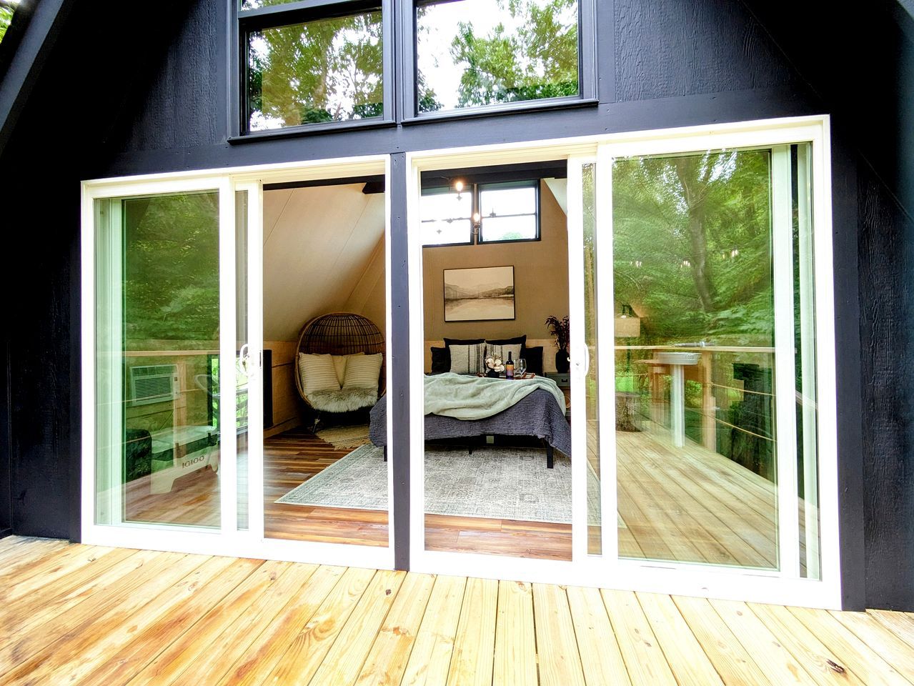 A modern bedroom with a large bed, a small table, and a chair. There is a sliding glass door leading to a balcony with a wooden deck.