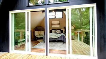 A modern bedroom with a large bed, a small table, and a chair. There is a sliding glass door leading to a balcony with a wooden deck.