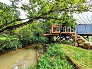 Een houten terras met een trap naar een huis, omringd door weelderige begroeiing en een rivier.