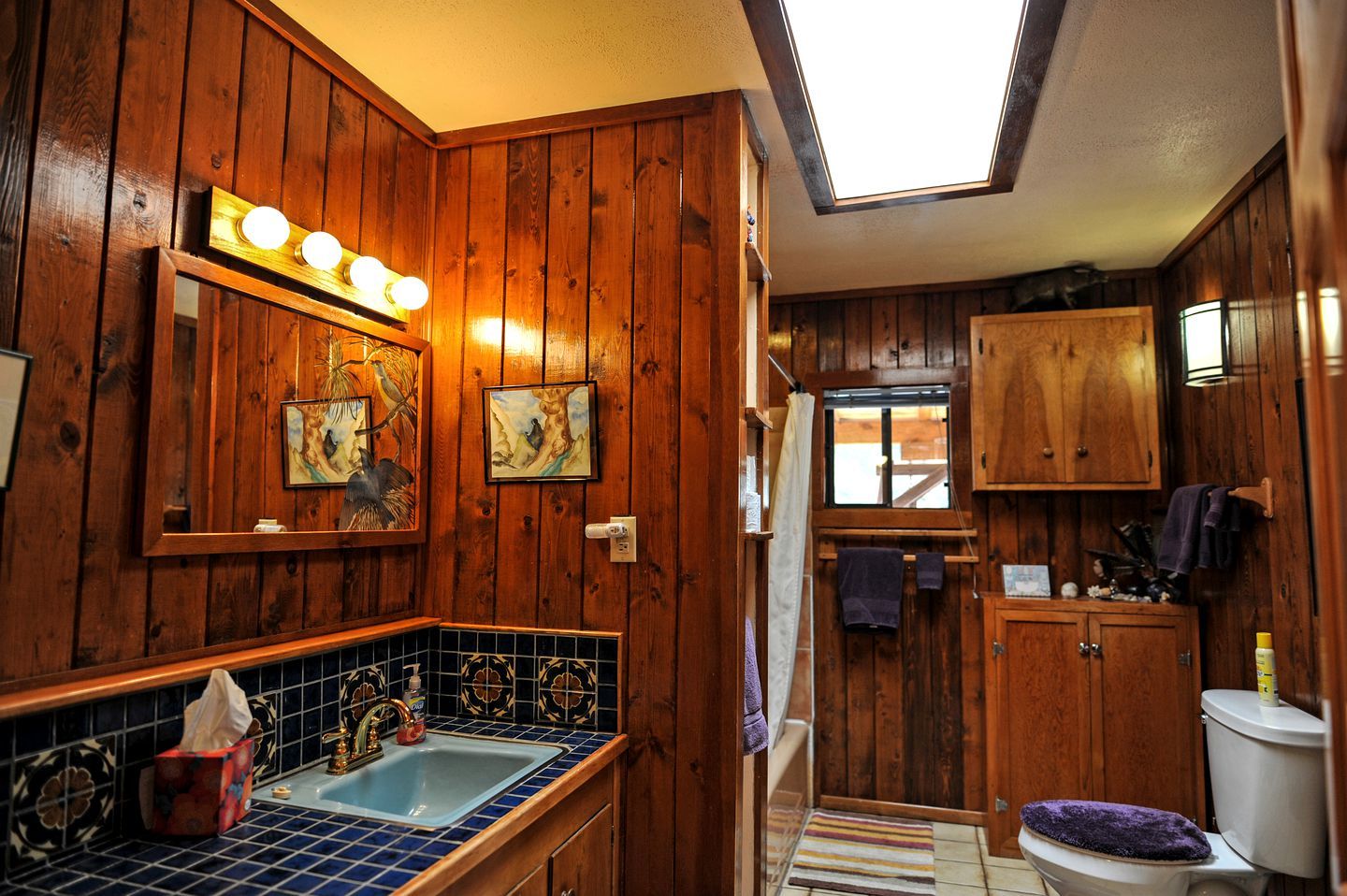 A compact, wood-paneled bathroom with a skylight, a sink with blue tiles, and wooden cabinets.