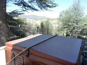 A hot tub with a wooden exterior and a metal frame, surrounded by a fence and trees, with a mountainous landscape in the background.