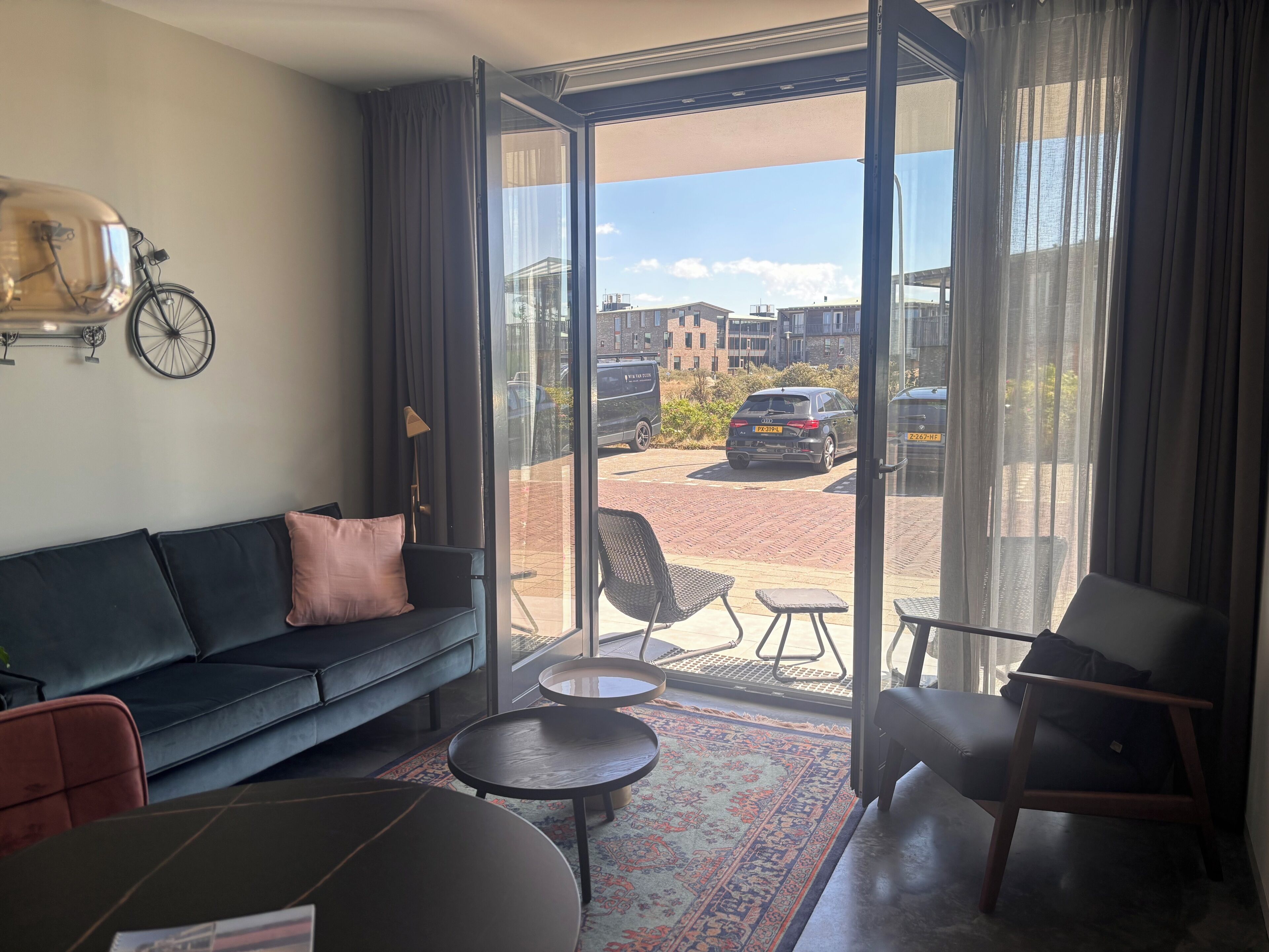 Un salon moderne avec un canapé, deux tables rondes, un tapis à motifs et une grande porte-fenêtre coulissante donnant sur un espace extérieur où l’on aperçoit une voiture et des bâtiments en arrière-plan.