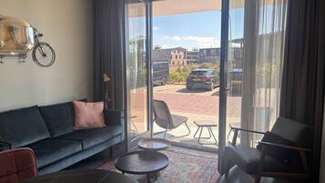 Un salon moderne avec un canapé, deux tables rondes, un tapis à motifs et une grande porte-fenêtre coulissante donnant sur un espace extérieur où l’on aperçoit une voiture et des bâtiments en arrière-plan.