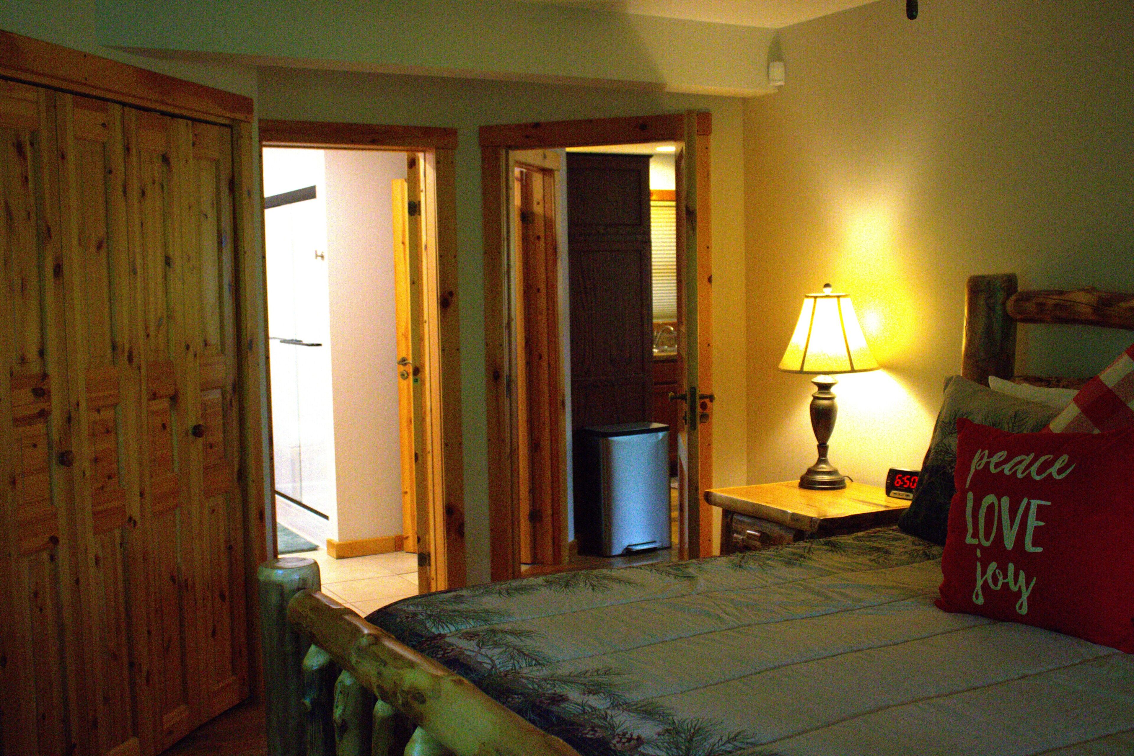 A bedroom with a bed, a nightstand with a lamp, a red pillow with 'peace LOVE joy' text, and wooden doors.