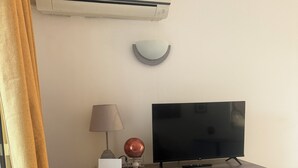 Une pièce avec un téléviseur à écran plat posé sur un meuble en bois, un climatiseur fixé au mur, une lampe et une fenêtre avec des rideaux.