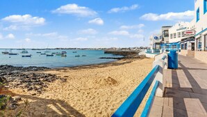 A beach with boats, a pier, and buildings with shops. - B-rent Bluemarlin Boutique Sea View – Central Apartment with Stunning Ocean Views (Corralejo)