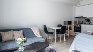 A modern living space with a sofa, a coffee table with a vase of flowers, a dining area with chairs, and a kitchenette with appliances.