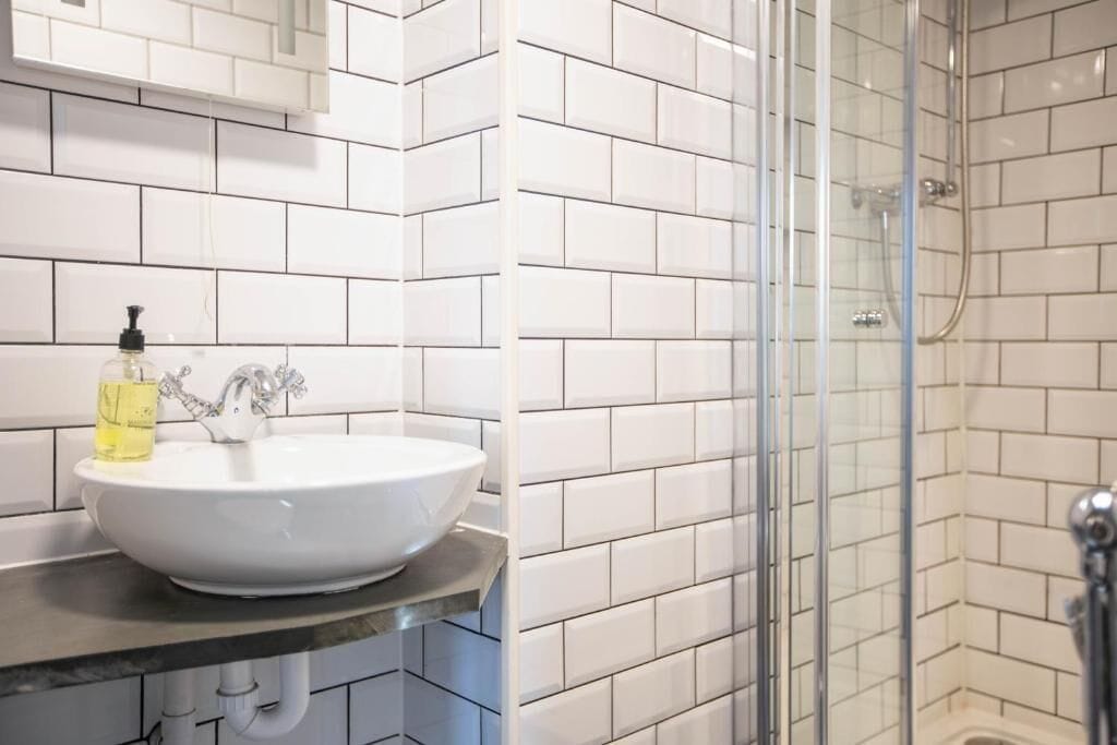 A bathroom with white tiled walls, a white sink with a soap dispenser, and a glass shower enclosure.