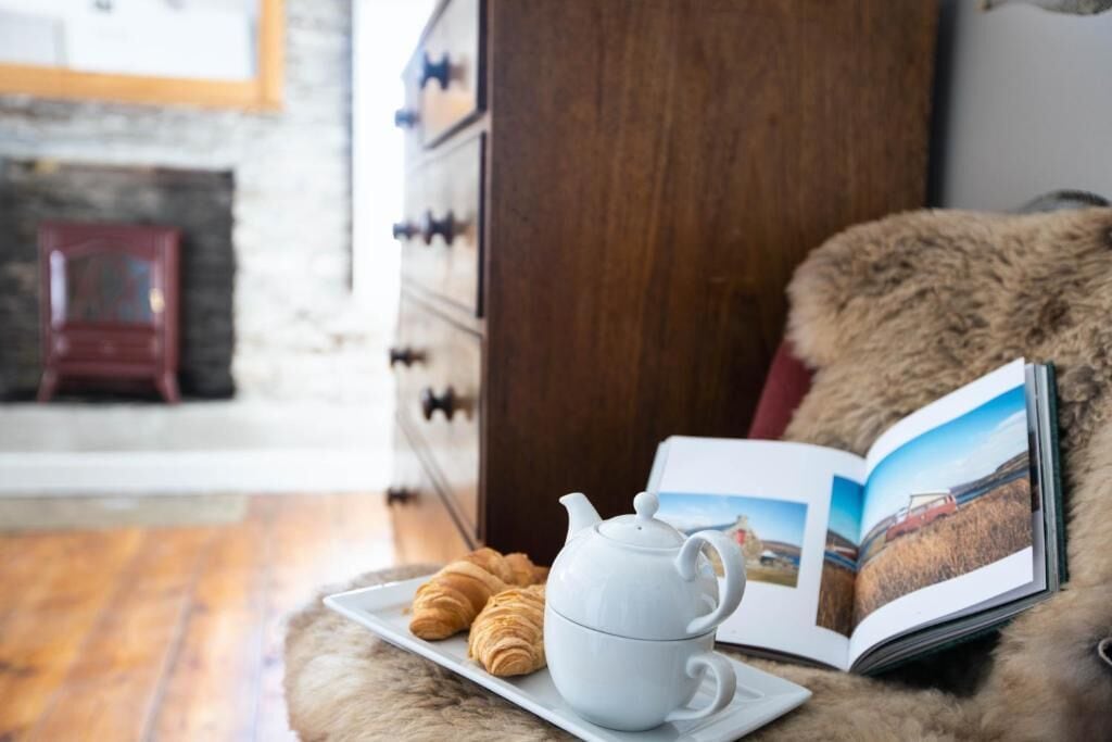 Una habitación acogedora con un aparador de madera, una tetera blanca sobre una bandeja con croissants, y un libro abierto con una fotografía de un paisaje.
