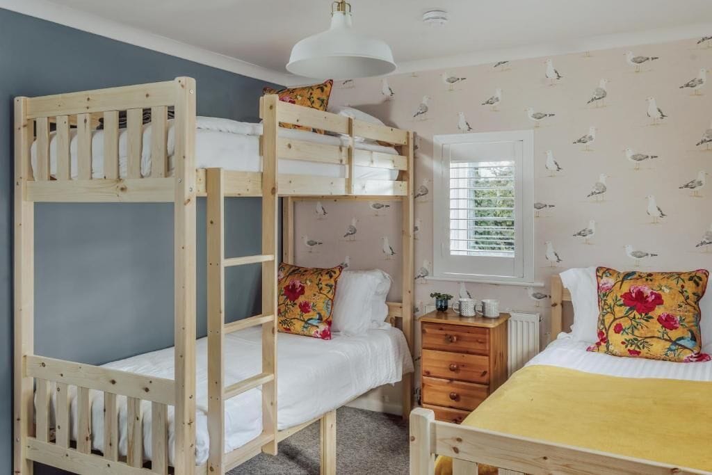 Una habitación con literas, un aparador, una ventana con persianas y un papel tapiz de gaviotas.